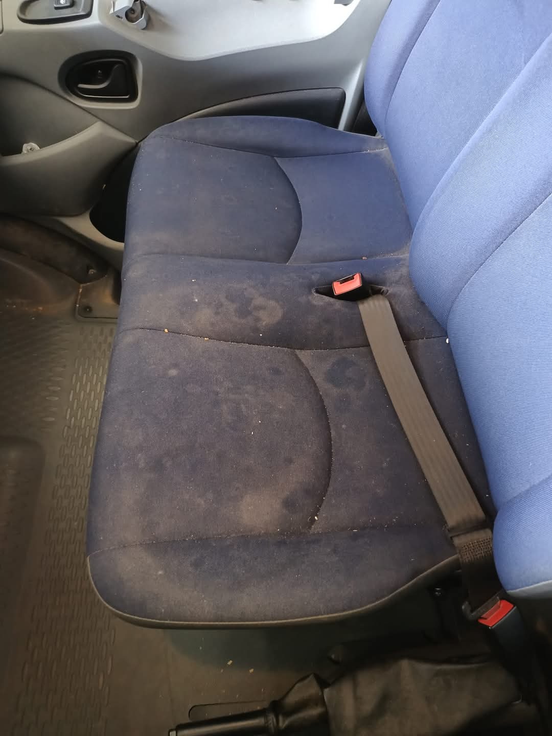 Brudne, niebieskie siedzenie samochodowe z plamami i zapiętym pasem bezpieczeństwa, widoczne elementy wnętrza pojazdu.