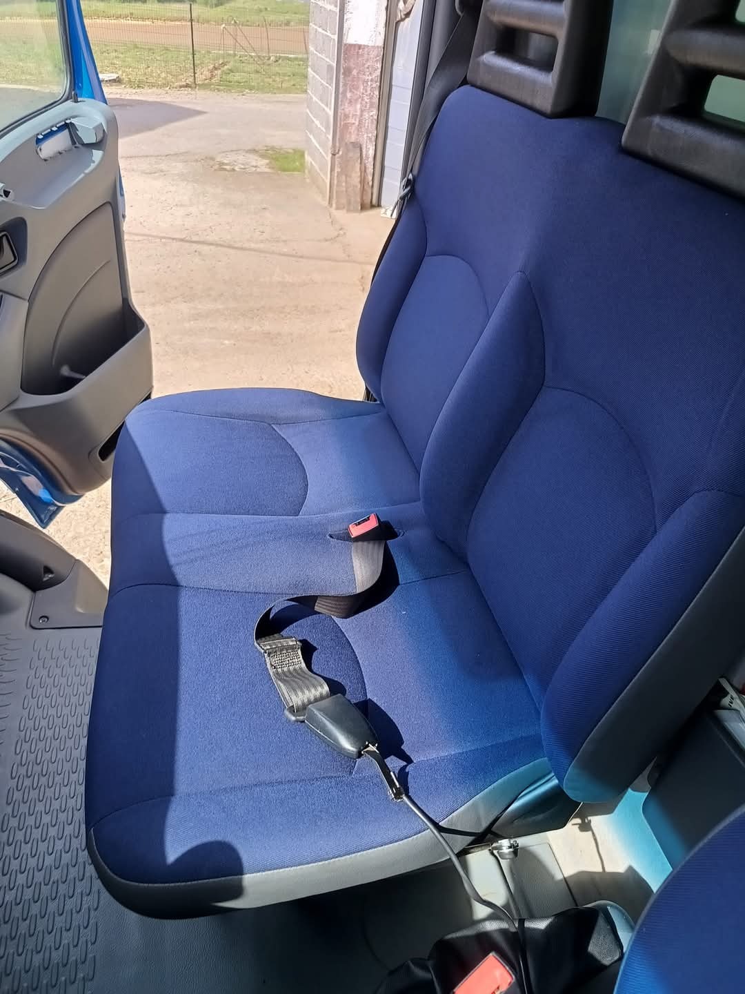 Wnętrze samochodu dostawczego: dwuosobowe siedzenie z niebieską tapicerką, widoczny pas bezpieczeństwa i otwarte drzwi kabiny.
