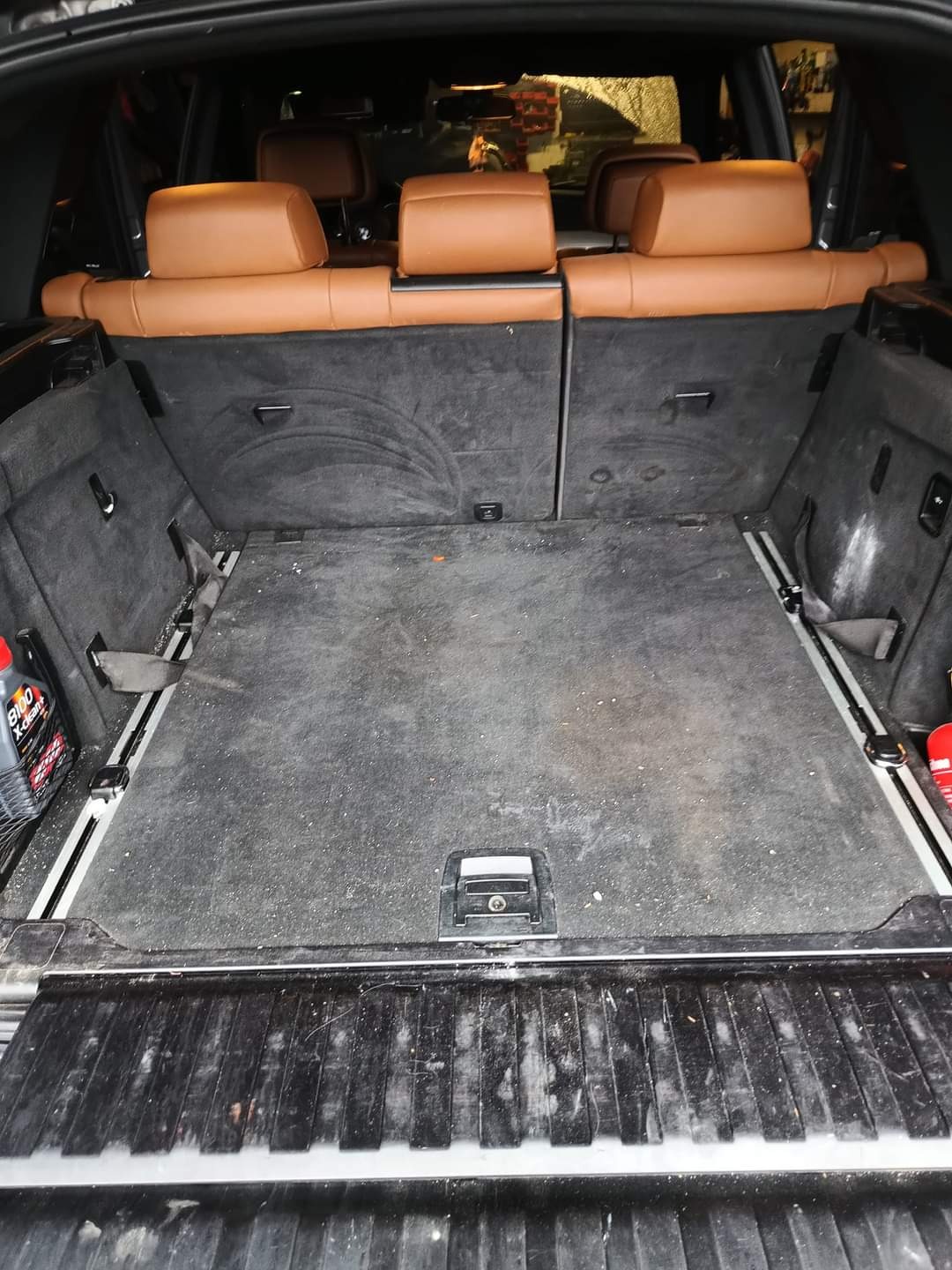 Wnętrze bagażnika SUV-a przed czyszczeniem, widoczny brud na tapicerce podłogi i ściankach bocznych, z butelką środka czyszczącego po lewej stronie.