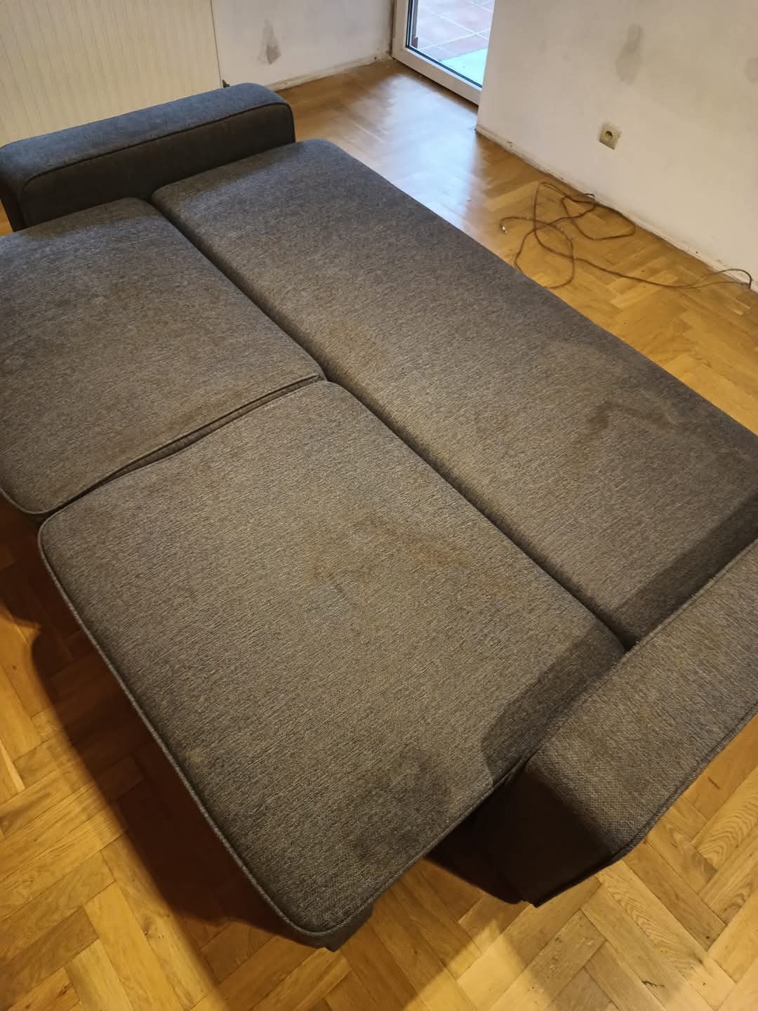 Szara, modułowa kanapa z widocznymi plamami na siedzisku, ustawiona na parkiecie w pokoju z białymi ścianami.