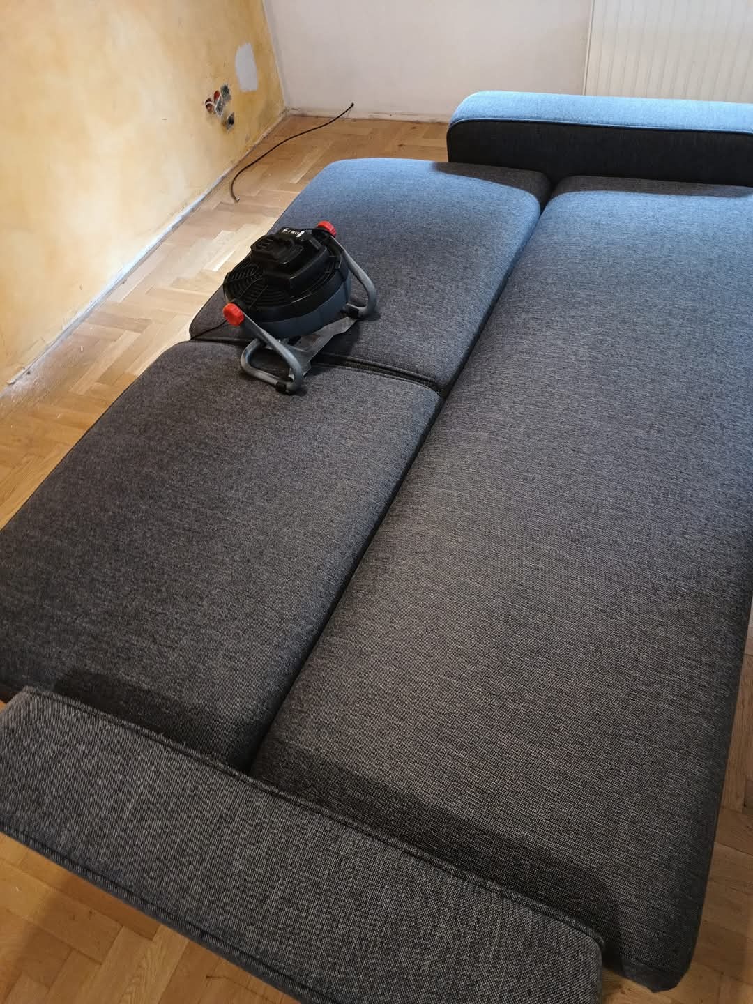 Szara, modułowa sofa z odkurzaczem piorącym na jednym z siedzisk, widoczne ślady prac remontowych na ścianie.