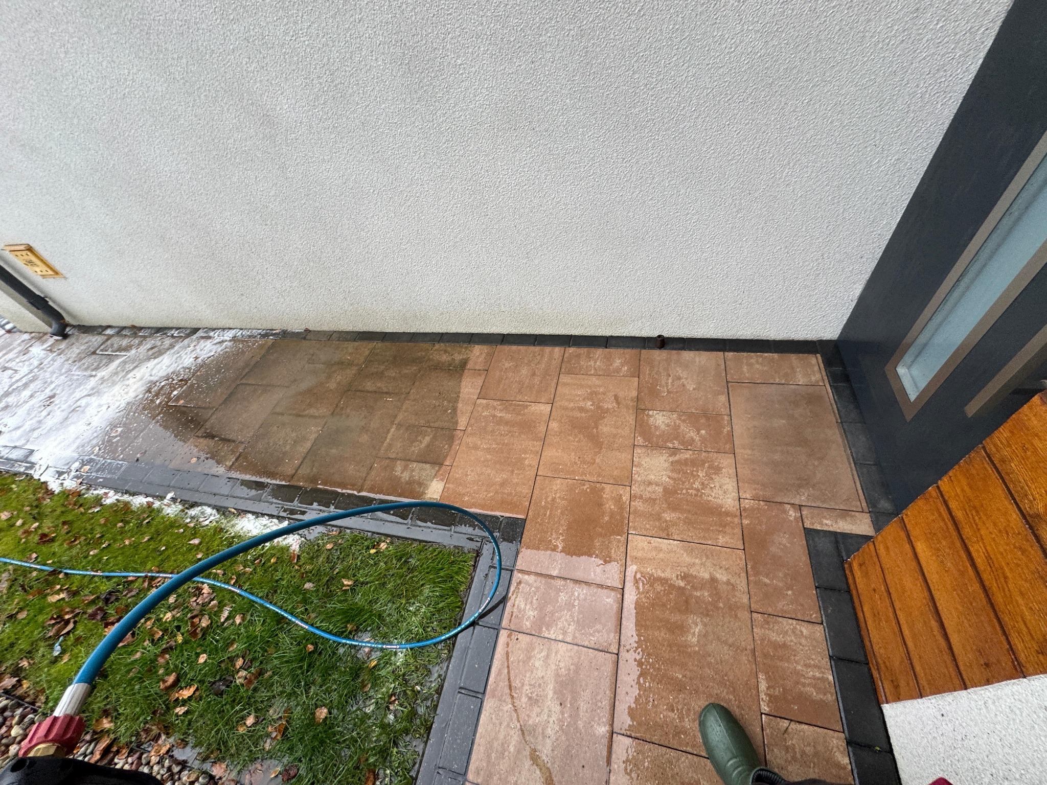 Częściowo umyta kostka brukowa przed budynkiem, widoczny wąż ciśnieniowy i trawnik z liśćmi.