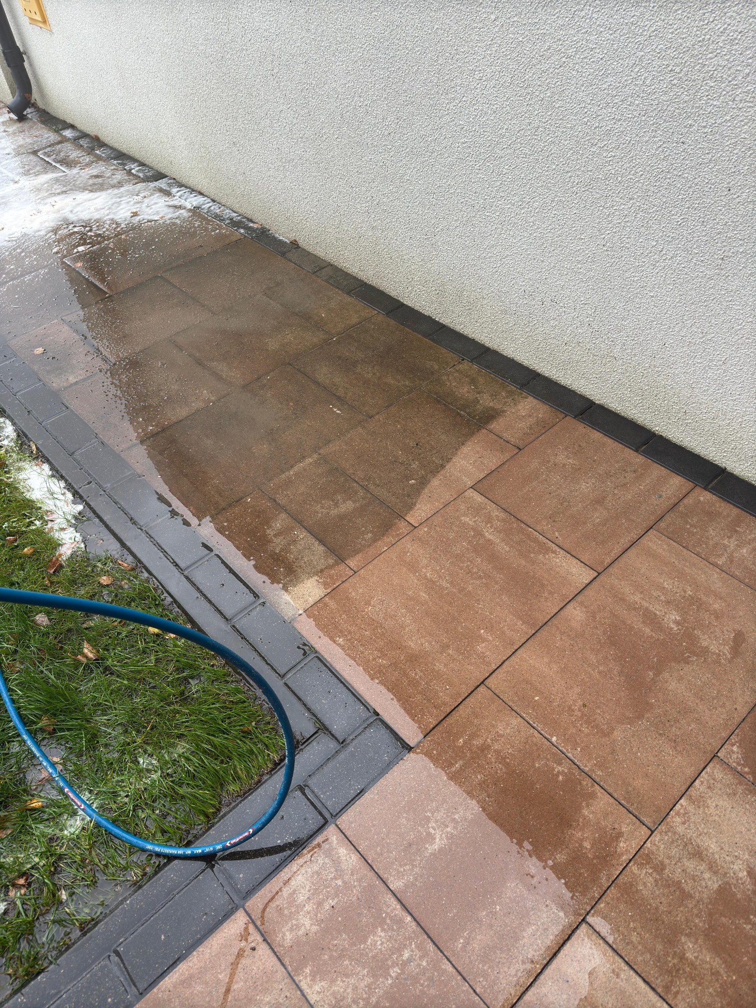 Częściowo umyta, brązowa kostka brukowa przylegająca do ściany budynku, widoczny fragment niebieskiego węża ogrodowego i trawnik. Część kostki jest mokra i czysta, a część sucha i brudna.