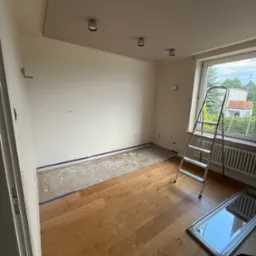 Pok&oacute;j w trakcie prac wykończeniowych: podłoga częściowo wyłożona panelami, ściany przygotowane do malowania, drabina przy oknie, listwy przypodłogowe zabezpieczone taśmą.