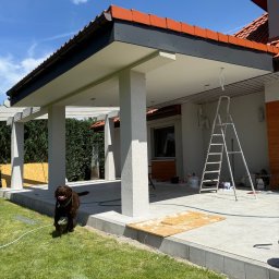 BRATAN MARCEL KONDRATOWICZ - Zadaszony taras w trakcie wykańczania z widoczną drabiną, narzędziami i biegnącym psem rasy labrador. Pomarańczowa dachówka i elementy drewniane.