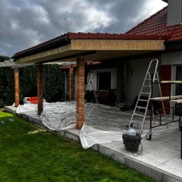 BRATAN MARCEL KONDRATOWICZ - Budowa zadaszenia tarasu przy domu jednorodzinnym. Widoczne ceglane słupy, drewniana konstrukcja dachu i rozłożona folia ochronna. Na tarasie drabiny i narzędzia.