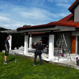 BRATAN MARCEL KONDRATOWICZ - Prace wykończeniowe na zewnątrz domu: Dwóch mężczyzn przy kolumnach, zabezpieczonych folią malarską, drabina oparta o ścianę. Widoczne narzędzia i akcesoria malarskie.