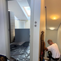 BRATAN MARCEL KONDRATOWICZ - Malarz w trakcie pracy przy schodach, zabezpieczone folią podłogi, widoczne narzędzia malarskie. Białe ściany i drewniana balustrada.