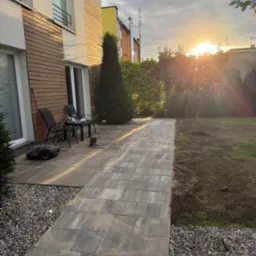 Ułożona kostka brukowa tworząca ścieżkę w ogrodzie, obok domu z drewnianą elewacją, w tle zachodzące słońce.