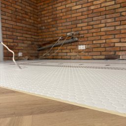 Biała mozaika heksagonalna na podłodze, kontrastująca z ceglaną ścianą w tle. Widoczne rury instalacji. Fragment ułożonych paneli podłogowych na pierwszym planie.