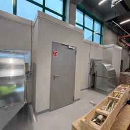 Wnętrze techniczne z szarymi ścianami, zielonymi ramami okiennymi i elementami instalacji wentylacyjnej. Na podłodze skrzynia z elementami montażowymi.