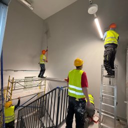 BRATAN MARCEL KONDRATOWICZ - Ekipa malarzy w kaskach i kamizelkach odblaskowych maluje klatkę schodową na szaro, używając wałków i rusztowań. Widoczne schody, lampy sufitowe i fragmenty instalacji.