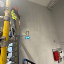 BRATAN MARCEL KONDRATOWICZ - Pracownik w kasku i kamizelce maluje ścianę na szaro z rusztowania. Widoczne zabezpieczenia przeciwpożarowe i schody. Ujęcie z dołu.