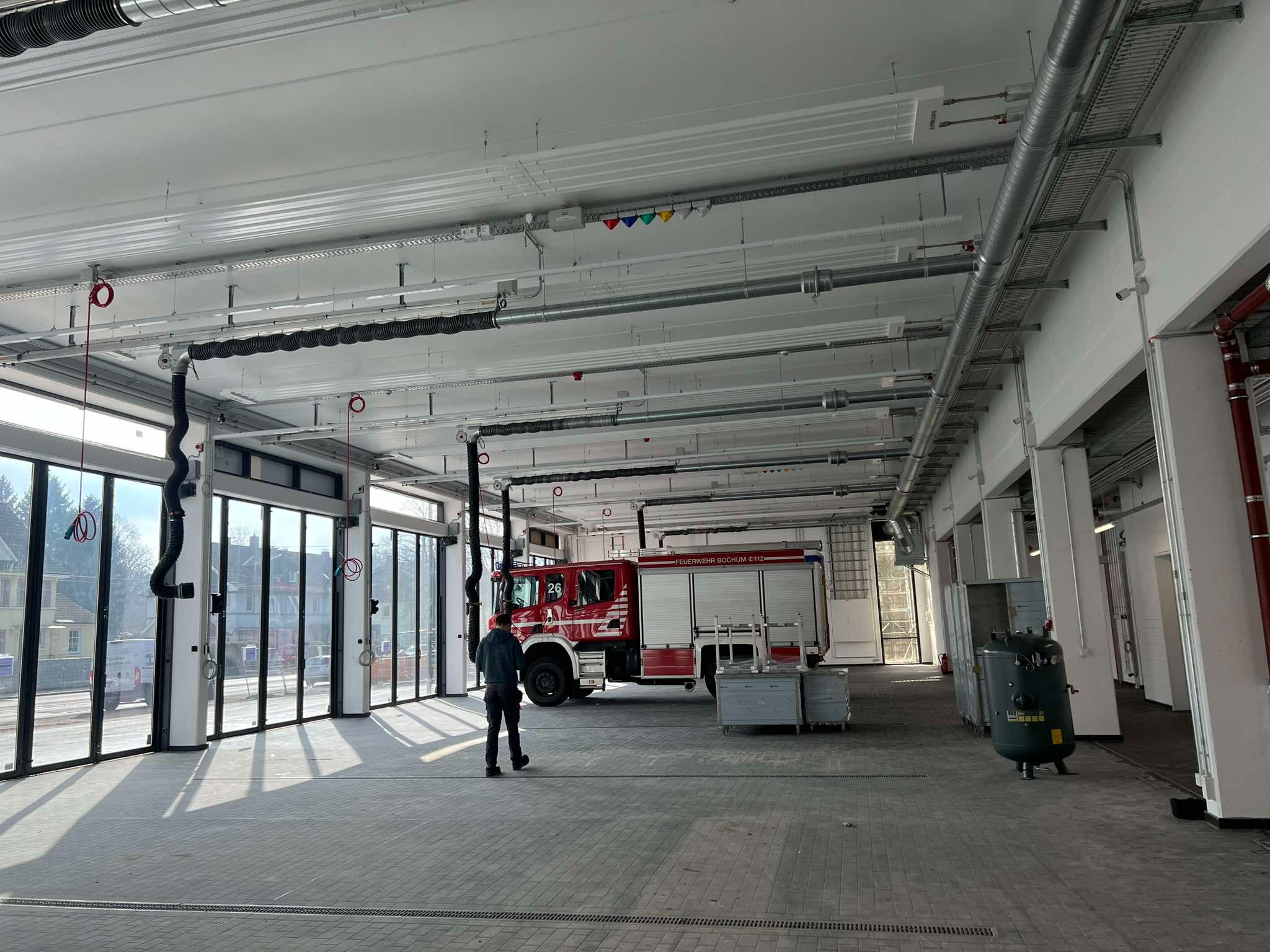 Wnętrze remizy strażackiej w Bochum z czerwonym wozem strażackim, widoczne instalacje pod sufitem i posadzka z szarej kostki brukowej.