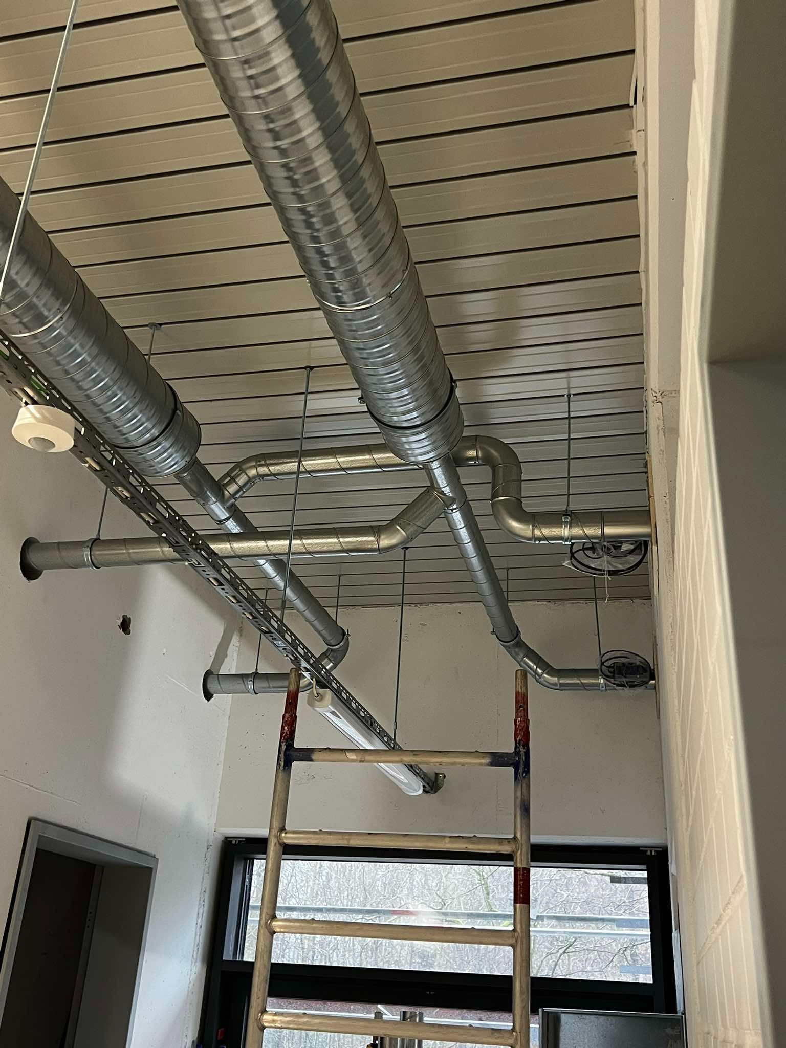 Widok z dołu na zamontowane, metalowe rury wentylacyjne pod sufitem z widoczną drabinką aluminiową.