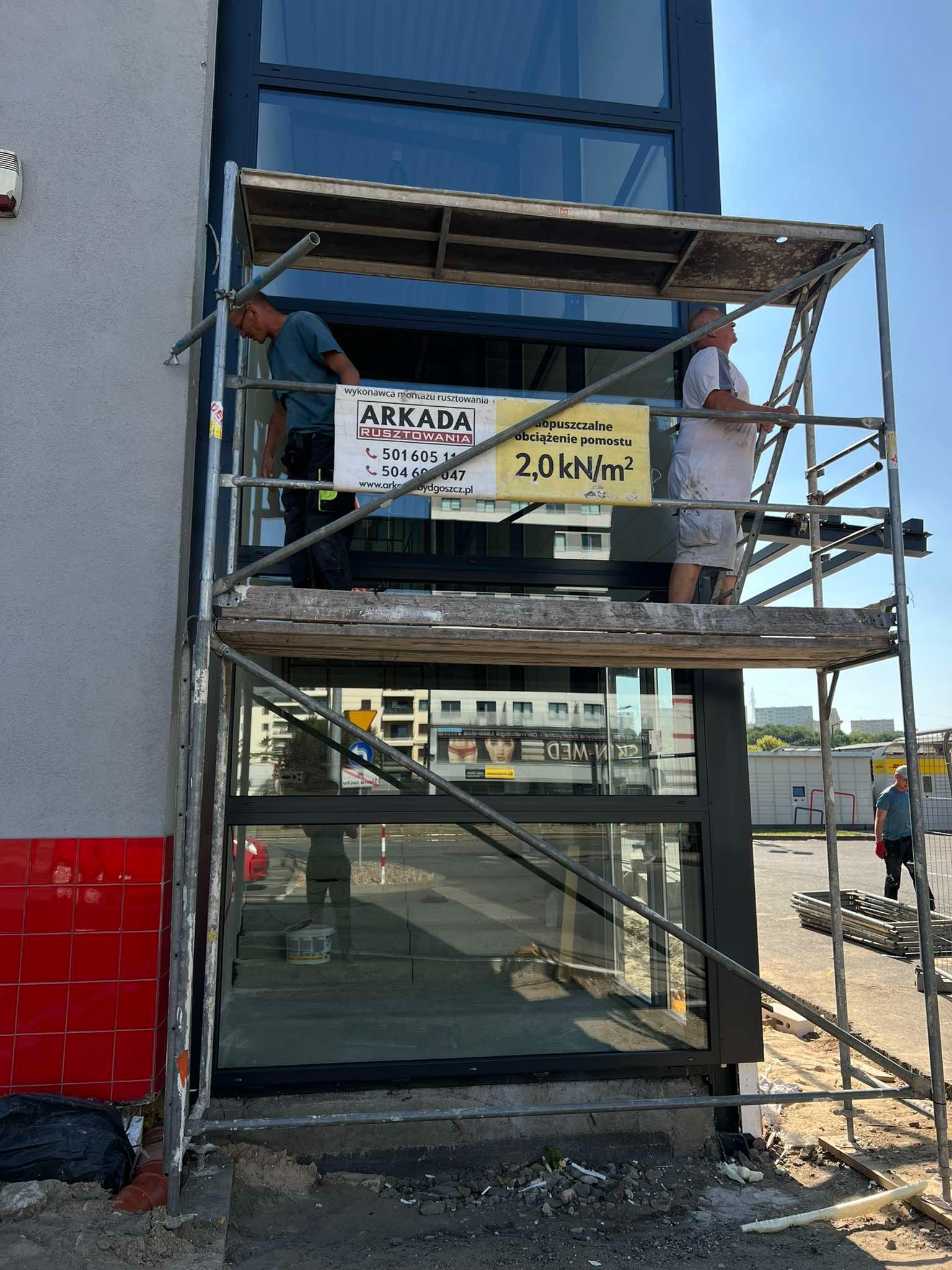 Praca na rusztowaniu przy fasadzie budynku Media Markt w Bydgoszczy, widoczni dwaj pracownicy na różnych poziomach rusztowania, tablica informacyjna o dopuszczalnym obciążeniu, czerwona płytka...