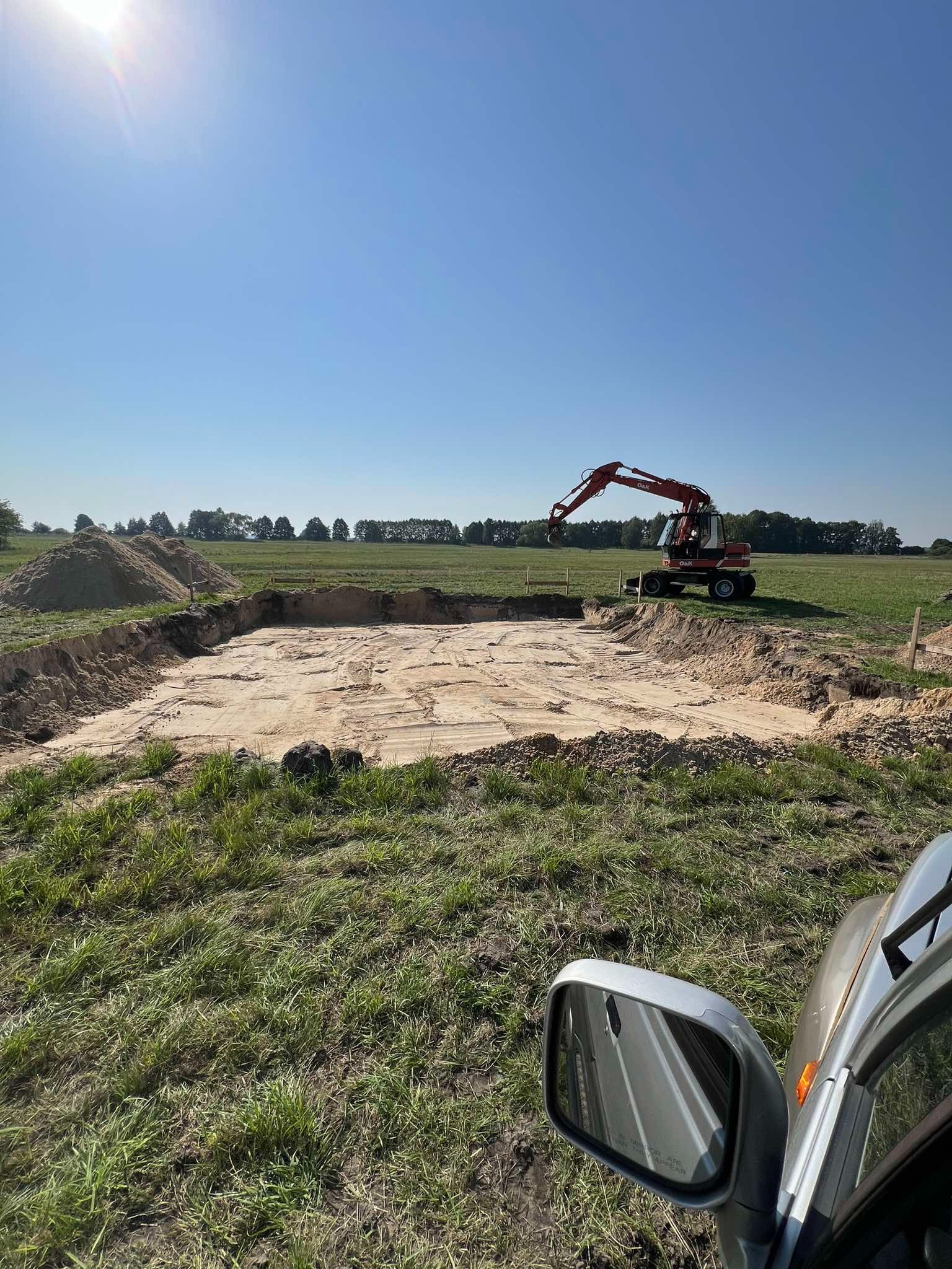 Wykonany wykop pod fundamenty budynku, widoczny zza lusterka samochodu, z czerwoną koparką w tle i stertami ziemi obok.