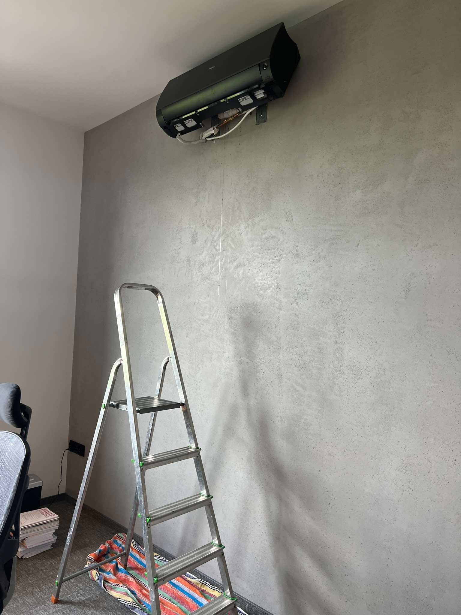 Montaż klimatyzacji: czarna jednostka wewnętrzna na szarej ścianie o fakturze betonu, widoczna drabina aluminiowa i wystające kable, etap instalacji.