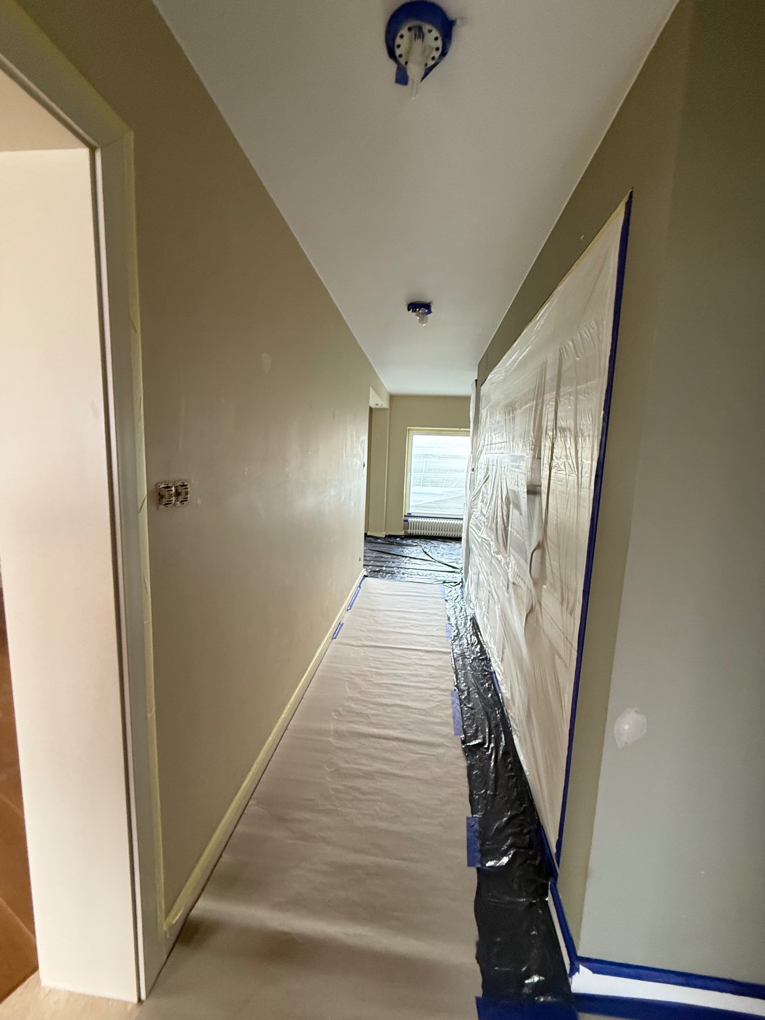 Korytarz w trakcie malowania: ściany w beżowym odcieniu, zabezpieczona folią podłoga i meble. Widoczne taśmy malarskie i zakryte lampy sufitowe.