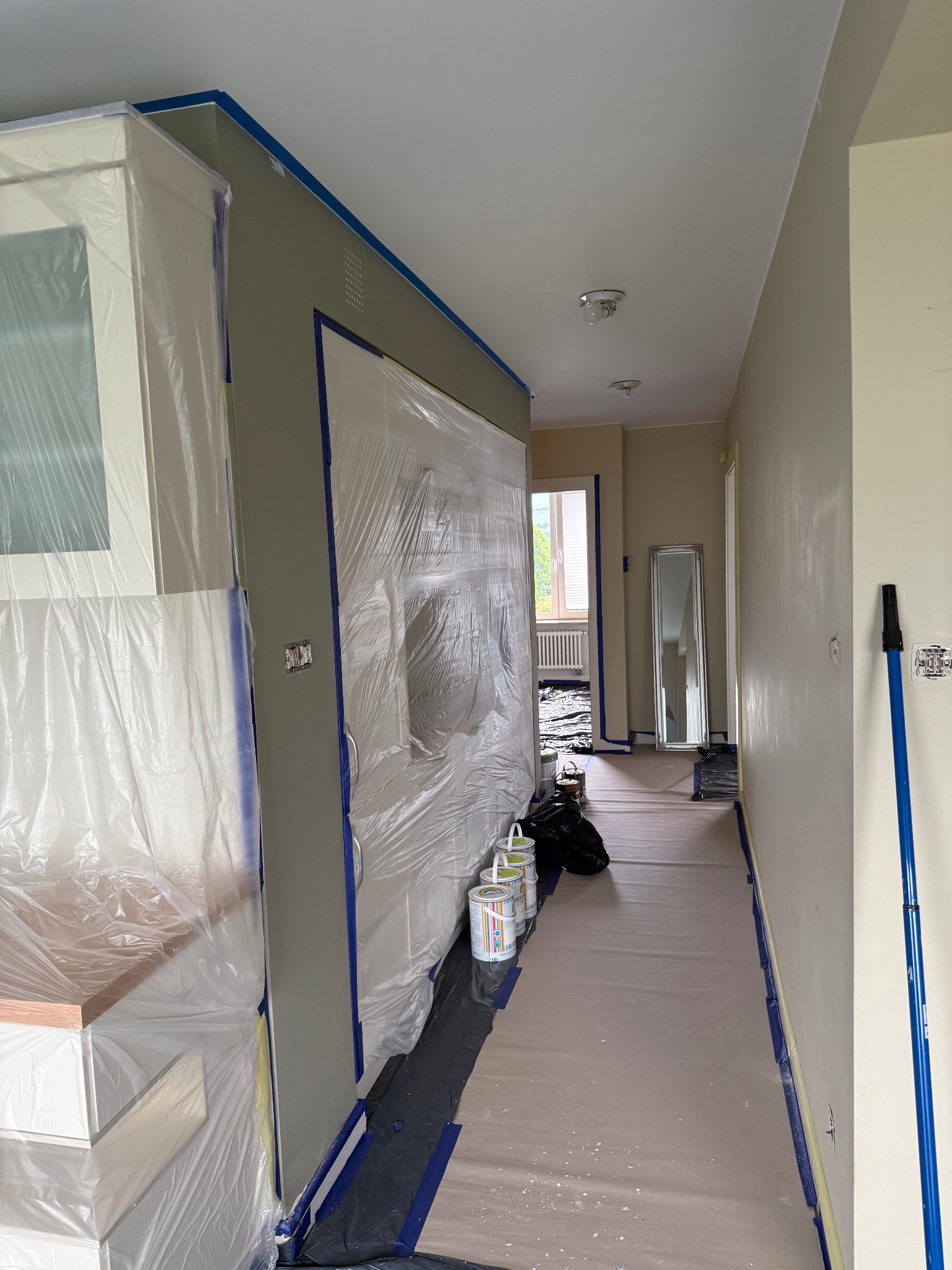 Przygotowanie do malowania: zabezpieczone meble folią, taśma malarska na ścianach i podłodze. Widoczne puszki z farbą i wałek malarski. Wnętrze w trakcie prac malarskich.