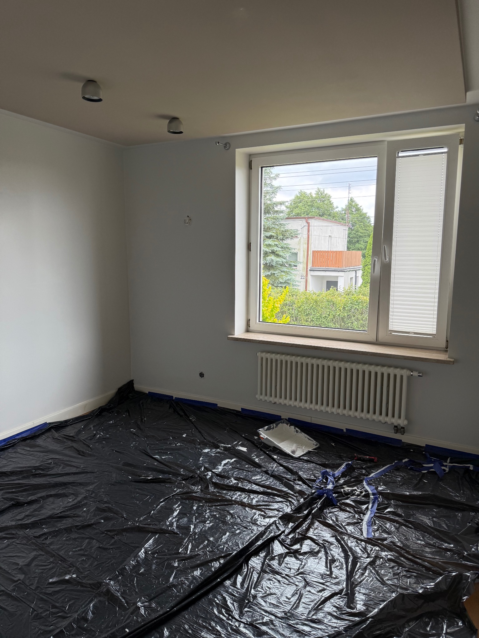 Przygotowanie do malowania: pokój zabezpieczony folią malarską, widoczny grzejnik pod oknem i puszka z farbą. Świeżo pomalowane ściany.