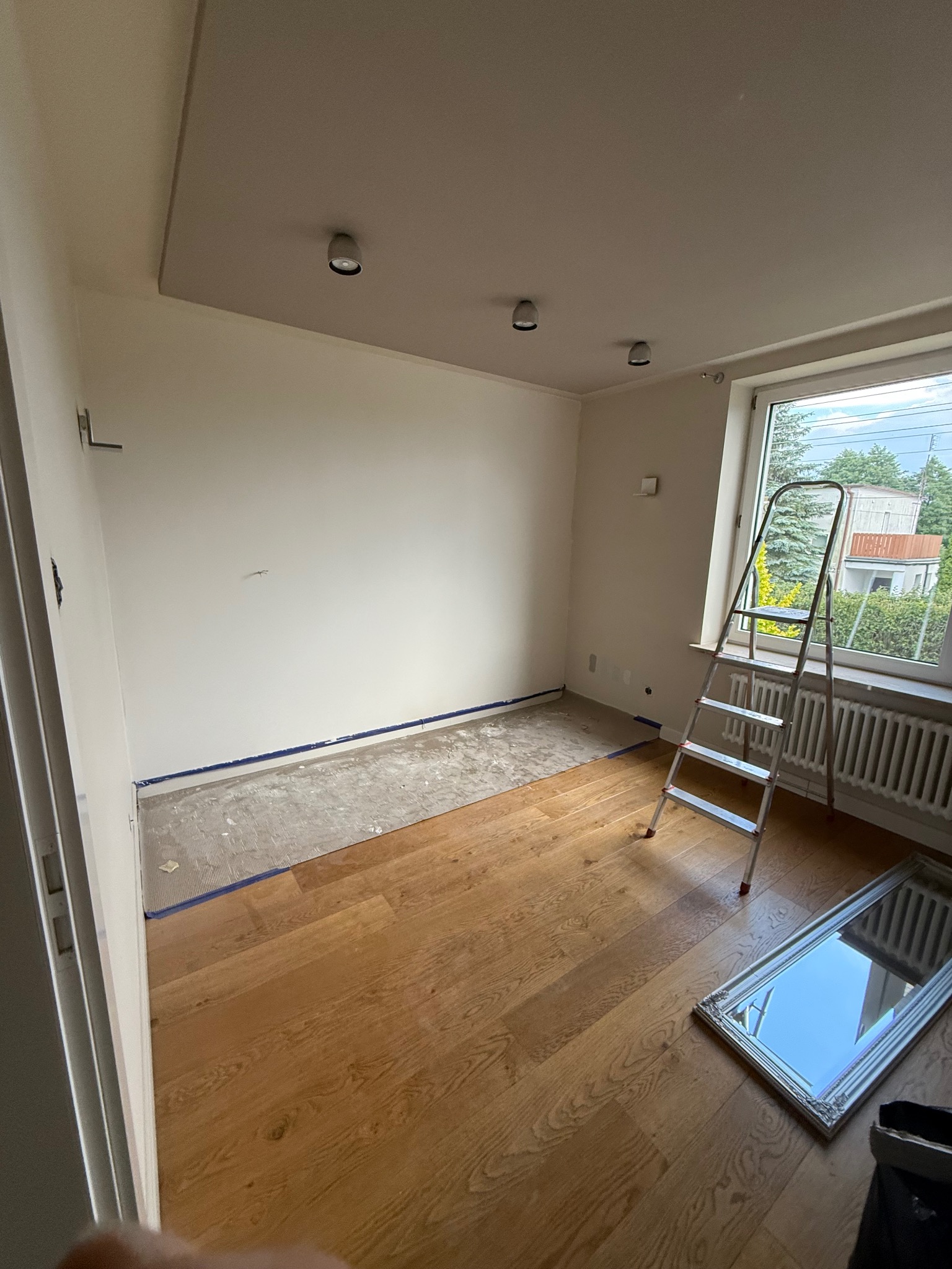 Pokój w trakcie prac wykończeniowych: podłoga częściowo wyłożona panelami, ściany przygotowane do malowania, drabina przy oknie, listwy przypodłogowe zabezpieczone taśmą.