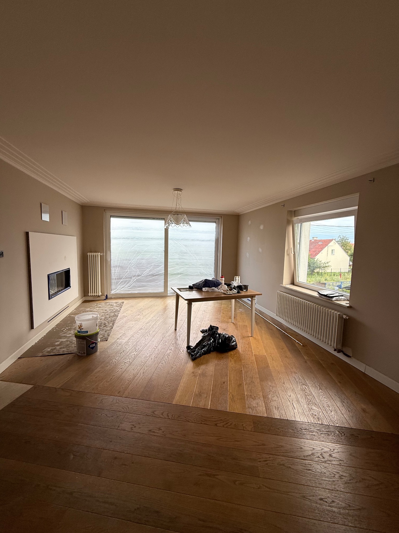 Pokój w trakcie malowania: ściany w beżowym odcieniu, okno zabezpieczone folią, widoczne pędzle, wiaderka z farbą i parkiet. Przygotowania do malowania mieszkania.