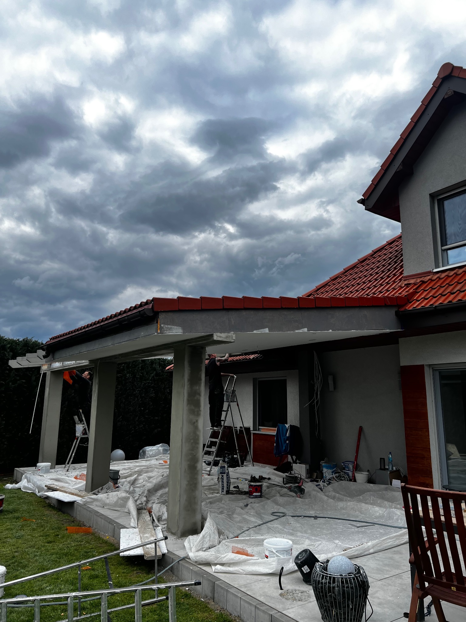 Dwóch mężczyzn na drabinach maluje podcienie domu z czerwonym dachem na tle zachmurzonego nieba. Teren zabezpieczony folią malarską, widoczne narzędzia i materiały budowlane.