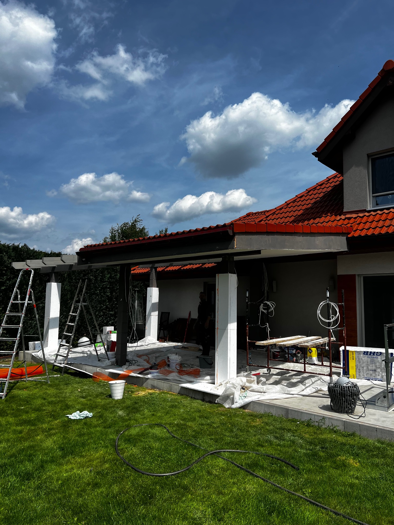 Prace wykończeniowe przy zadaszeniu tarasu domu jednorodzinnego. Widoczne rusztowania, drabiny, wiadro z farbą i zabezpieczenia. W tle budynek z czerwoną dachówką i błękitne niebo.