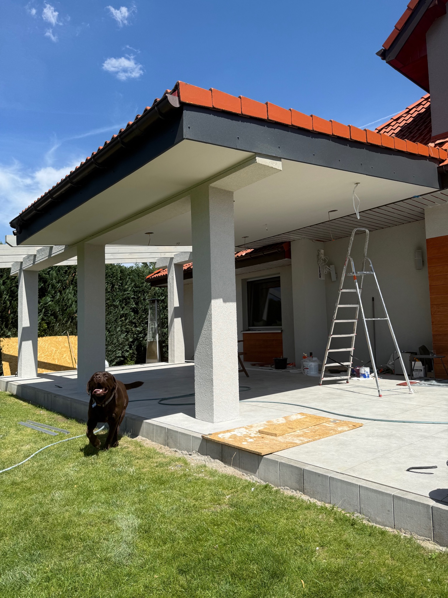 Zadaszony taras w trakcie wykańczania z widoczną drabiną, narzędziami i biegnącym psem rasy labrador. Pomarańczowa dachówka i elementy drewniane.