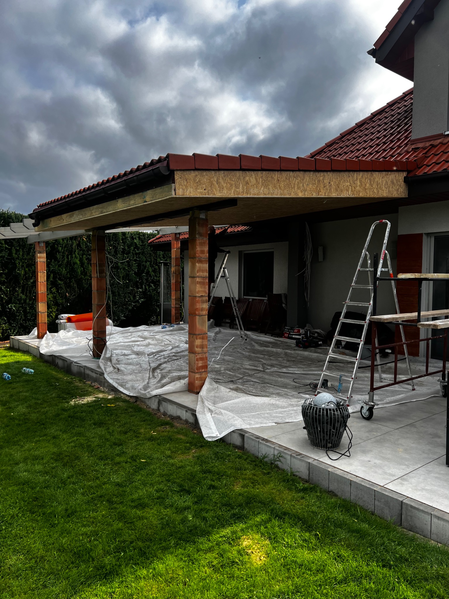 Budowa zadaszenia tarasu przy domu jednorodzinnym. Widoczne ceglane słupy, drewniana konstrukcja dachu i rozłożona folia ochronna. Na tarasie drabiny i narzędzia.
