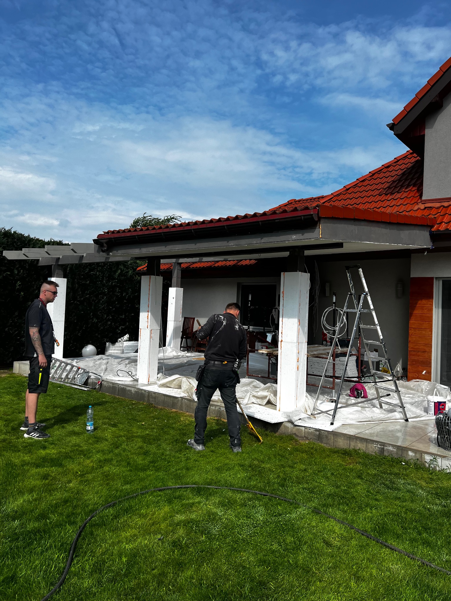 Prace wykończeniowe na zewnątrz domu: Dwóch mężczyzn przy kolumnach, zabezpieczonych folią malarską, drabina oparta o ścianę. Widoczne narzędzia i akcesoria malarskie.