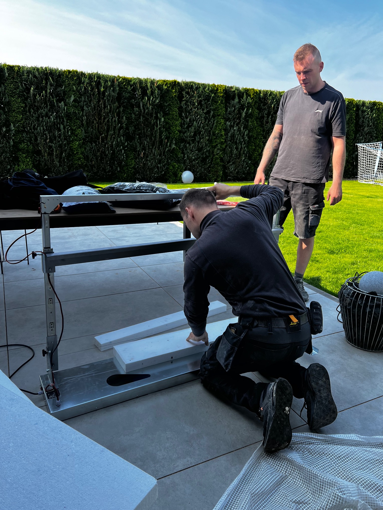 Dwóch mężczyzn przy pracy z urządzeniem do cięcia styropianu na zewnątrz, na tle zielonego trawnika i żywopłotu. Jeden klęczy, drugi stoi i nadzoruje.