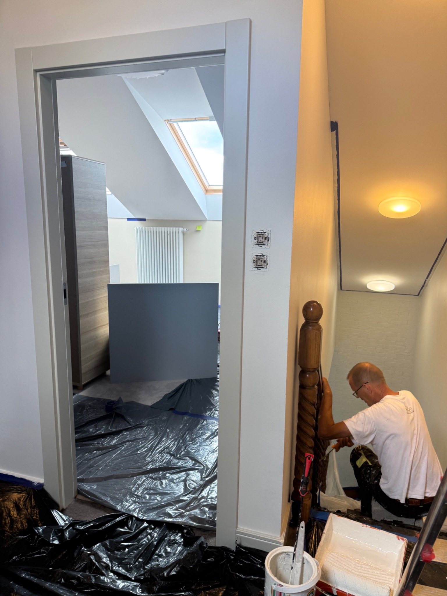 Malarz w trakcie pracy przy schodach, zabezpieczone folią podłogi, widoczne narzędzia malarskie. Białe ściany i drewniana balustrada.