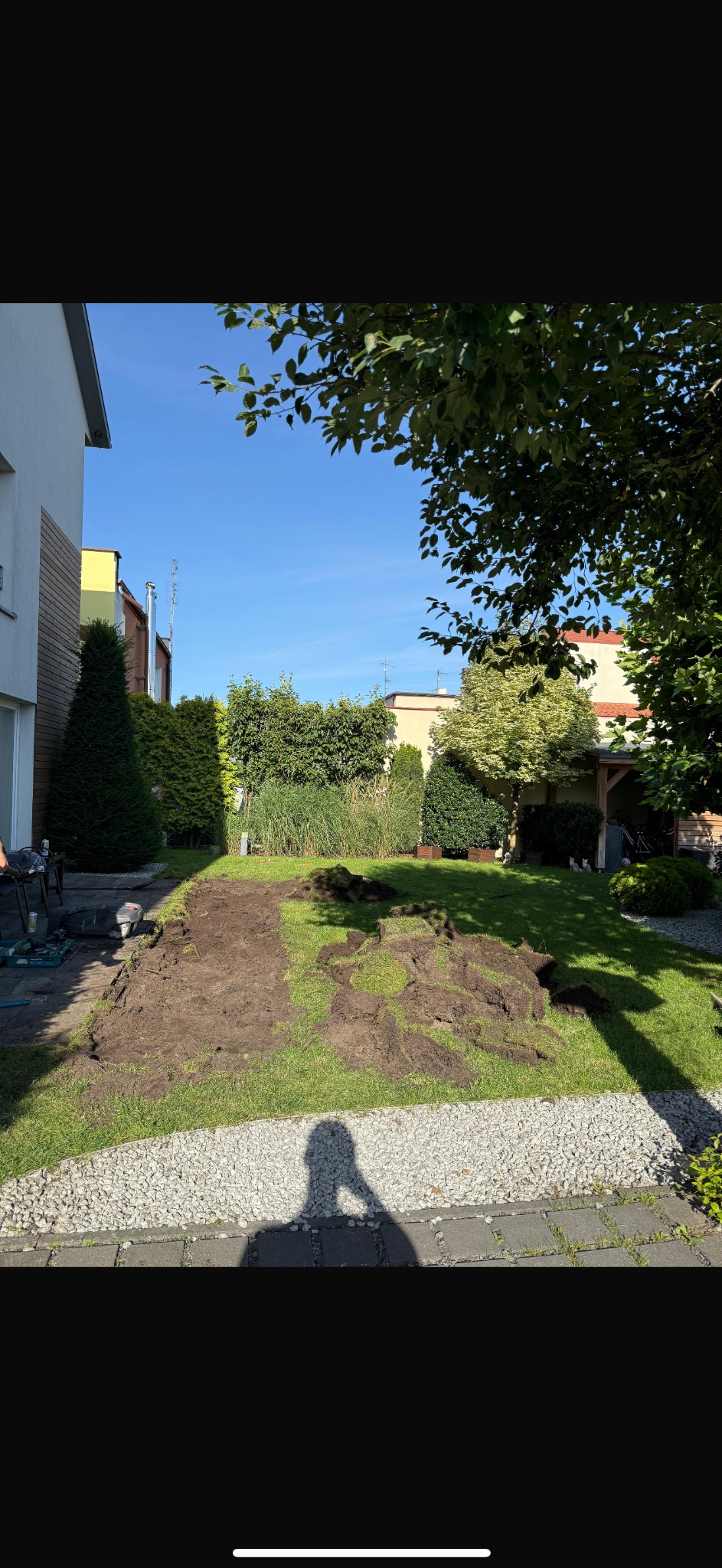 Fragment ogrodu z wykopanym obszarem, przygotowanym pod nasadzenia; widoczne narzędzia ogrodnicze i cień osoby robiącej zdjęcie.