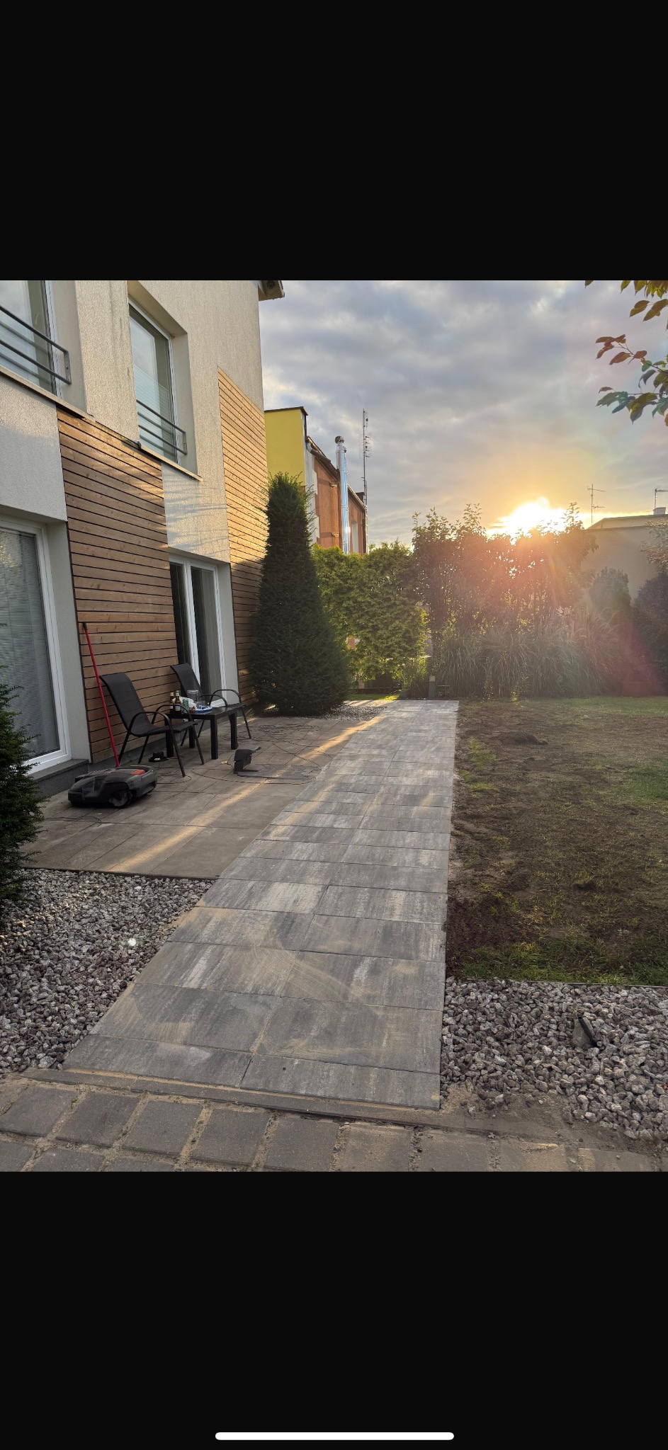 Ułożona kostka brukowa tworząca ścieżkę w ogrodzie, obok domu z drewnianą elewacją, w tle zachodzące słońce.