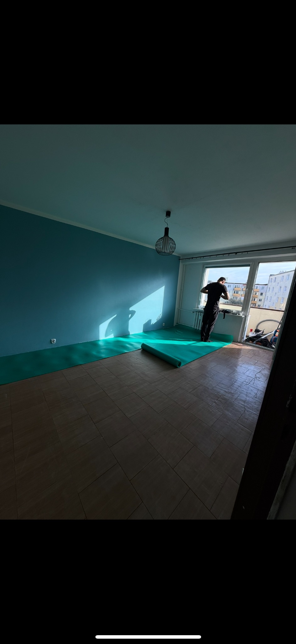 Przygotowanie do układania paneli: rozłożony podkład piankowy na brązowej podłodze, mężczyzna przy oknie, błękitne ściany, nowoczesna lampa.