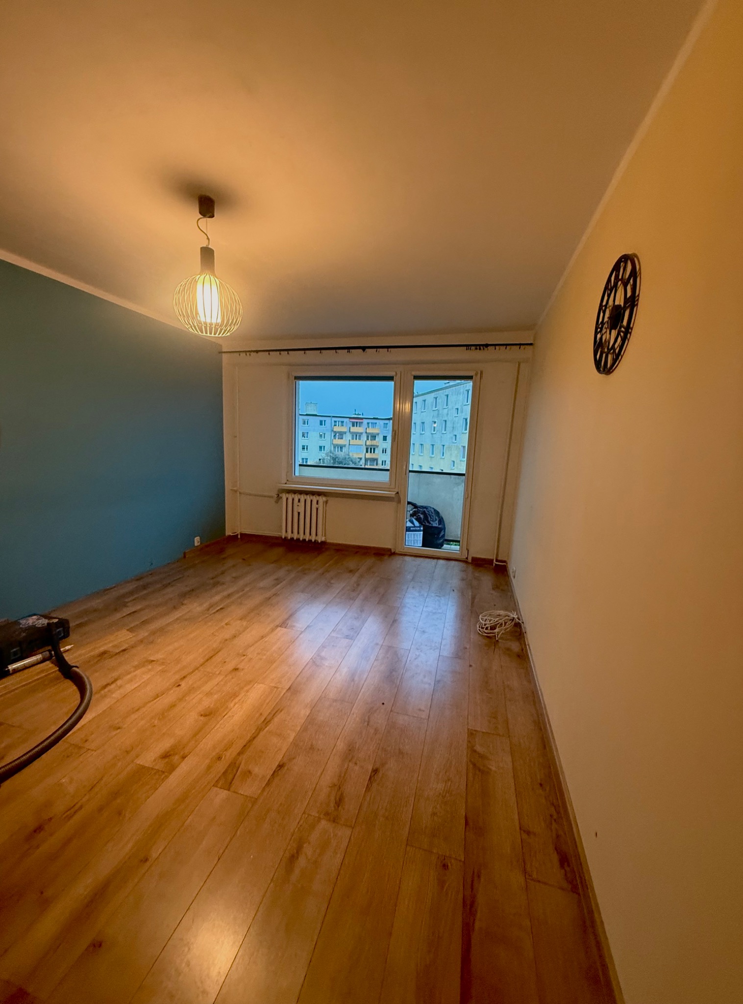 Pokój po remoncie: panele podłogowe, pomalowana na niebiesko ściana, okno balkonowe, zegar na ścianie. Widoczne narzędzia.