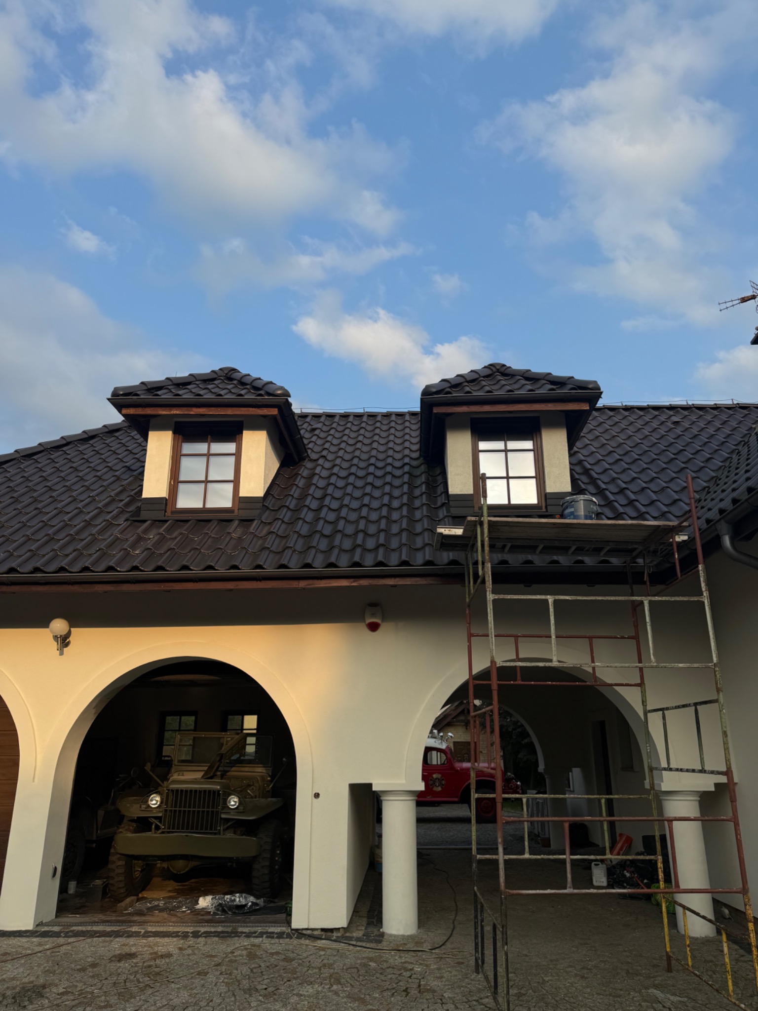 Elewacja domu z rusztowaniem przy oknie na poddaszu, widoczny fragment dachu z dachówką. W garażu zaparkowany wojskowy samochód.