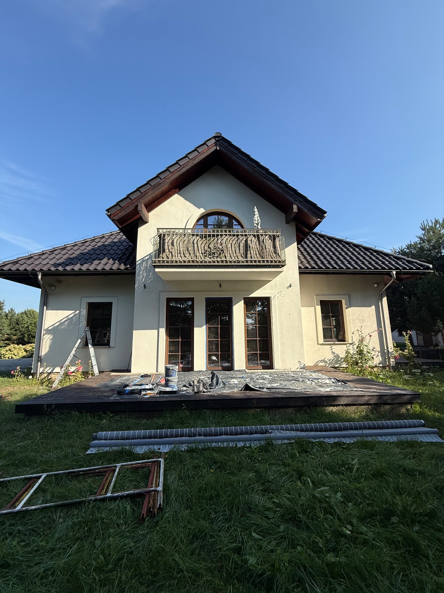 Dom dwupiętrowy z balkonem w trakcie prac wykończeniowych. Na tarasie farby i narzędzia malarskie, na trawniku leżą elementy rusztowania i rolki materiału.