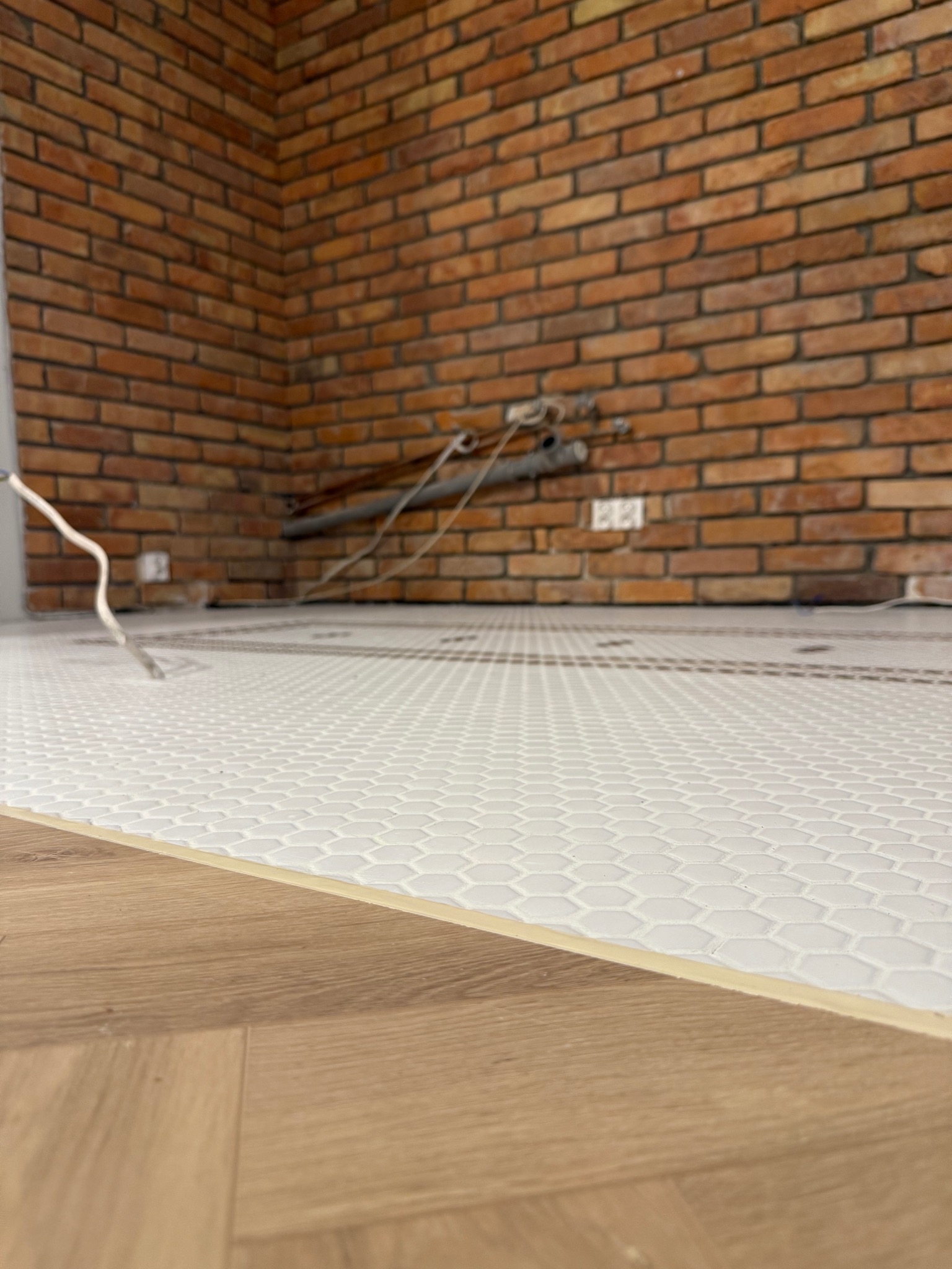 Biała mozaika heksagonalna na podłodze, kontrastująca z ceglaną ścianą w tle. Widoczne rury instalacji. Fragment ułożonych paneli podłogowych na pierwszym planie.