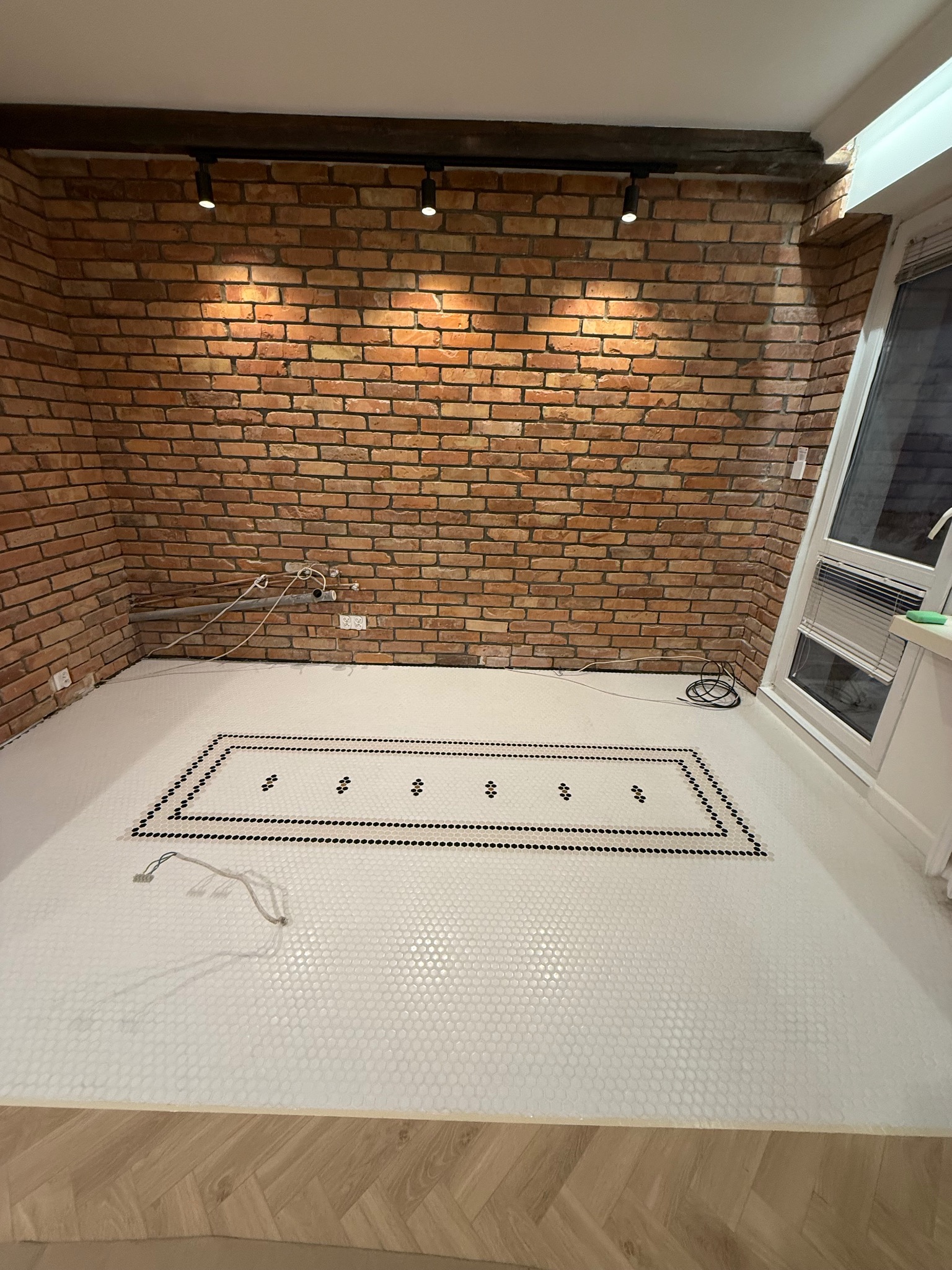 Wykończona podłoga z białej mozaiki z czarnym wzorem w pomieszczeniu z ceglaną ścianą i oknem. Widoczne elementy instalacji hydraulicznej i oświetlenie.