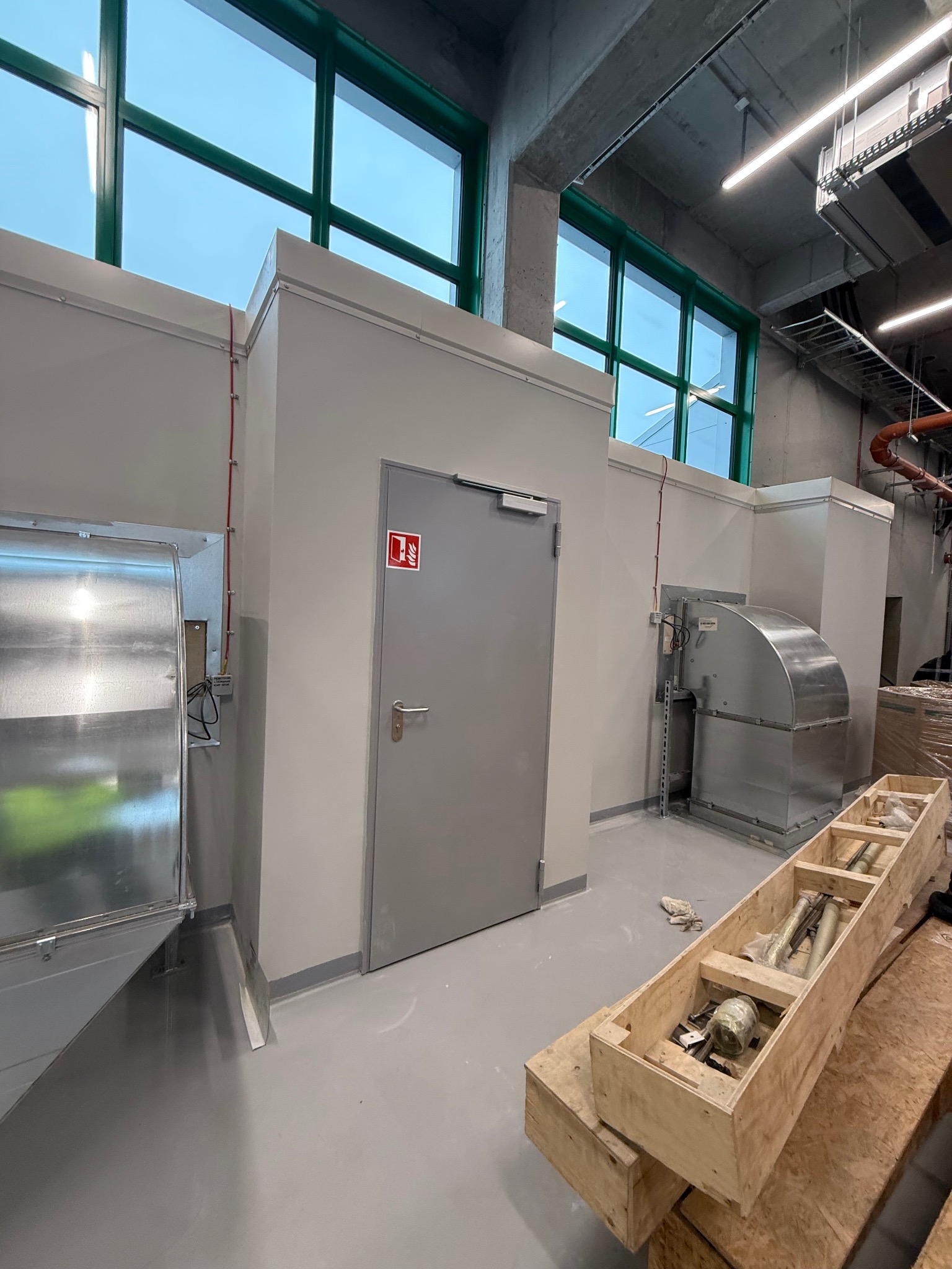 Wnętrze techniczne z szarymi ścianami, zielonymi ramami okiennymi i elementami instalacji wentylacyjnej. Na podłodze skrzynia z elementami montażowymi.