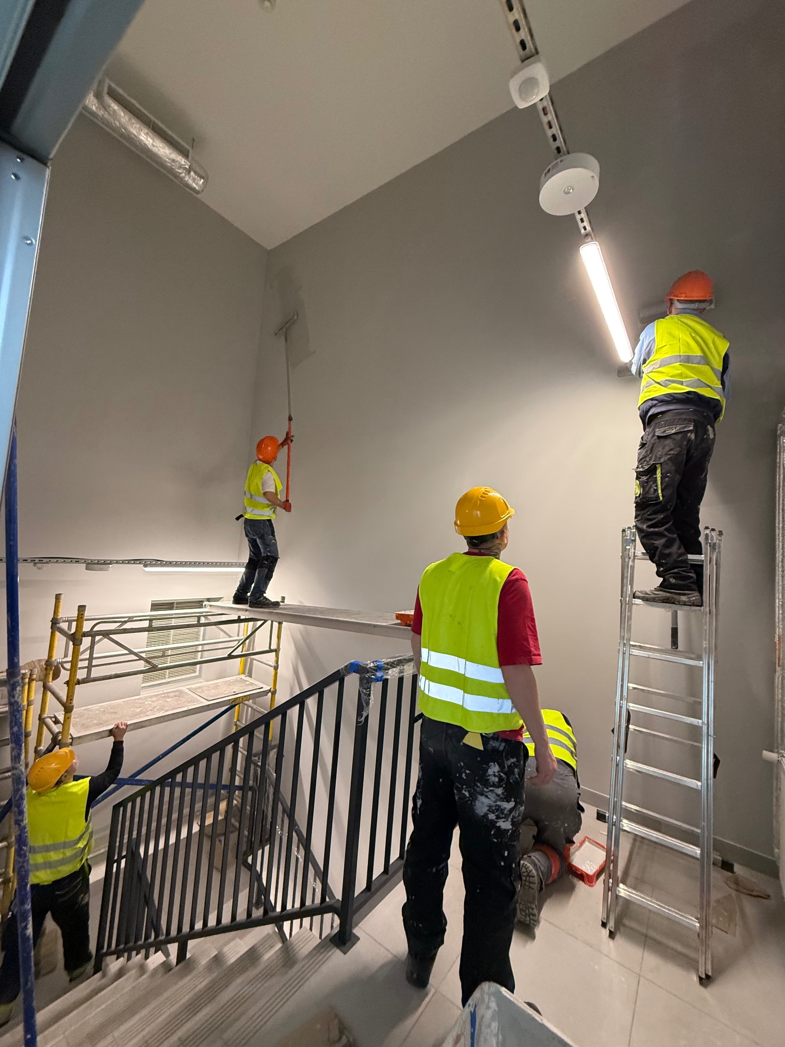 Ekipa malarzy w kaskach i kamizelkach odblaskowych maluje klatkę schodową na szaro, używając wałków i rusztowań. Widoczne schody, lampy sufitowe i fragmenty instalacji.