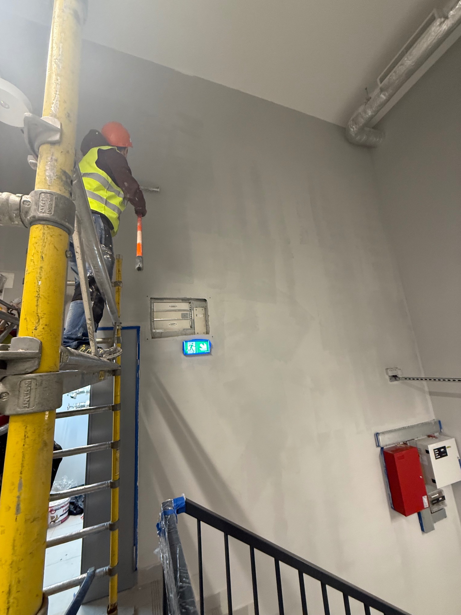 Pracownik w kasku i kamizelce maluje ścianę na szaro z rusztowania. Widoczne zabezpieczenia przeciwpożarowe i schody. Ujęcie z dołu.