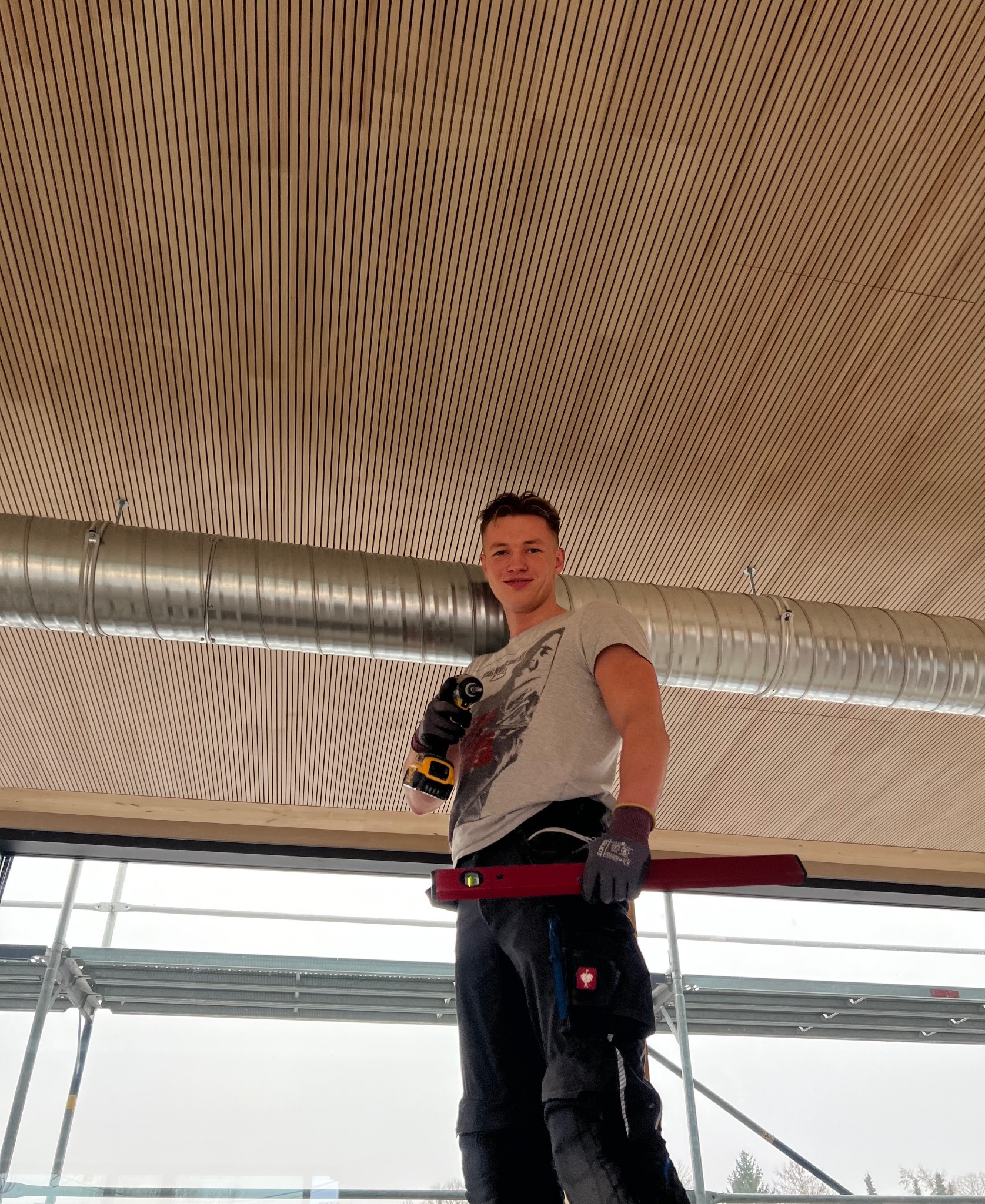 Pracownik z wiertarką i poziomicą na tle drewnianego sufitu i instalacji wentylacyjnej w Bydgoszczy. Widok z dołu, rusztowanie w tle.