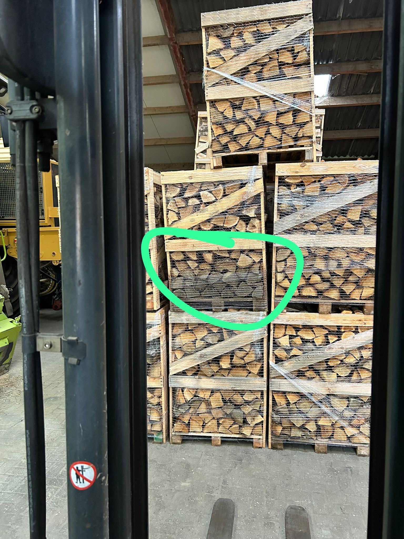 Stosy palet z pociętym drewnem, owinięte folią, widoczne przez konstrukcję wózka widłowego, z zaznaczoną paletą z ciemniejszym drewnem.