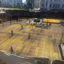Zbrojenie fundamentu z żółtą folią, widok z góry na plac budowy z pracownikami w odblaskowych ubraniach. Metalowe ścianki oporowe.