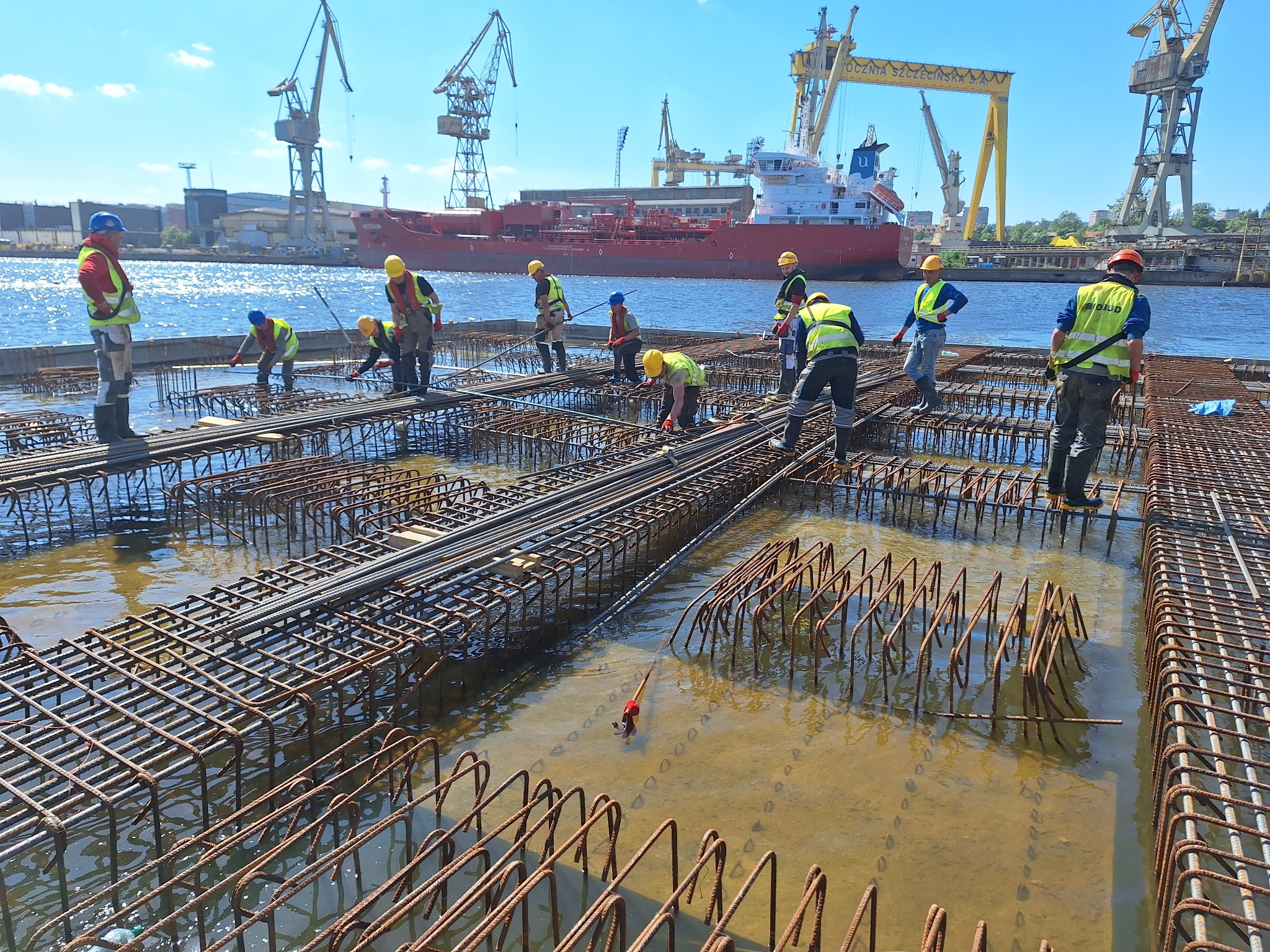 Pracownicy w odblaskowych kamizelkach i kaskach montują zbrojenie na terenie budowy w wodzie, w tle dźwigi portowe i czerwony statek.