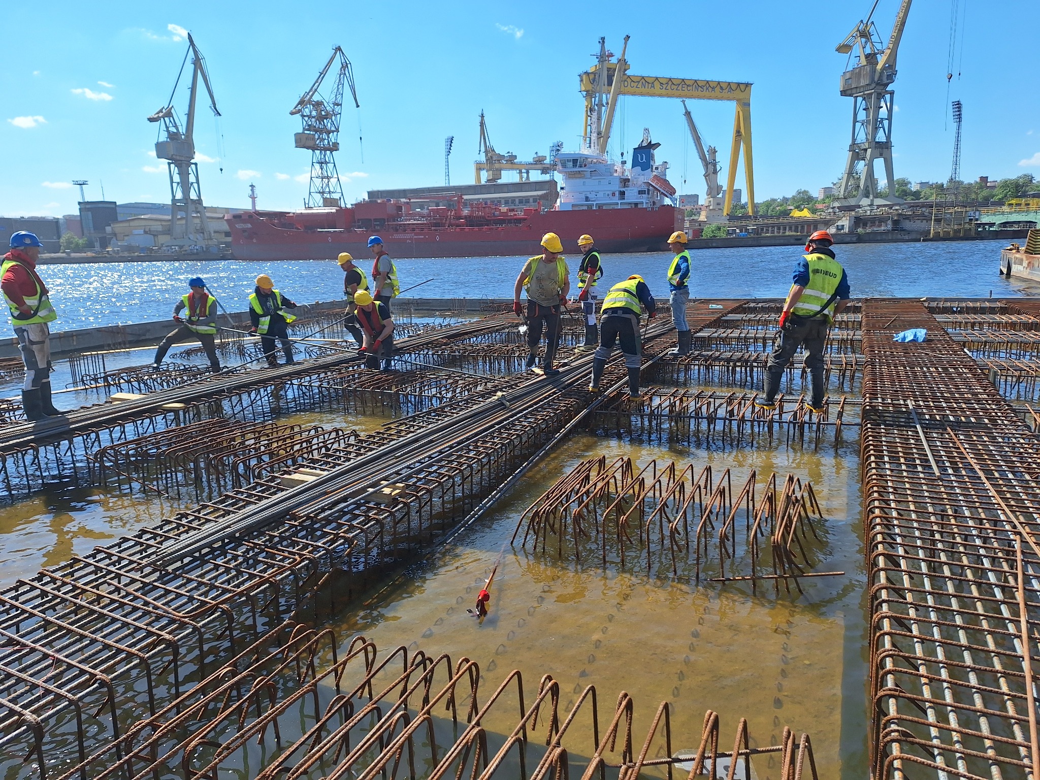 Zbrojarze pracują nad konstrukcją zbrojeniową zalewaną wodą, na tle stoczni z dźwigami i czerwonym statkiem.